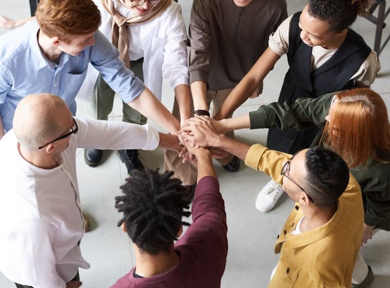 A diverse group of professionals joins hands in a team-building exercise indoors.