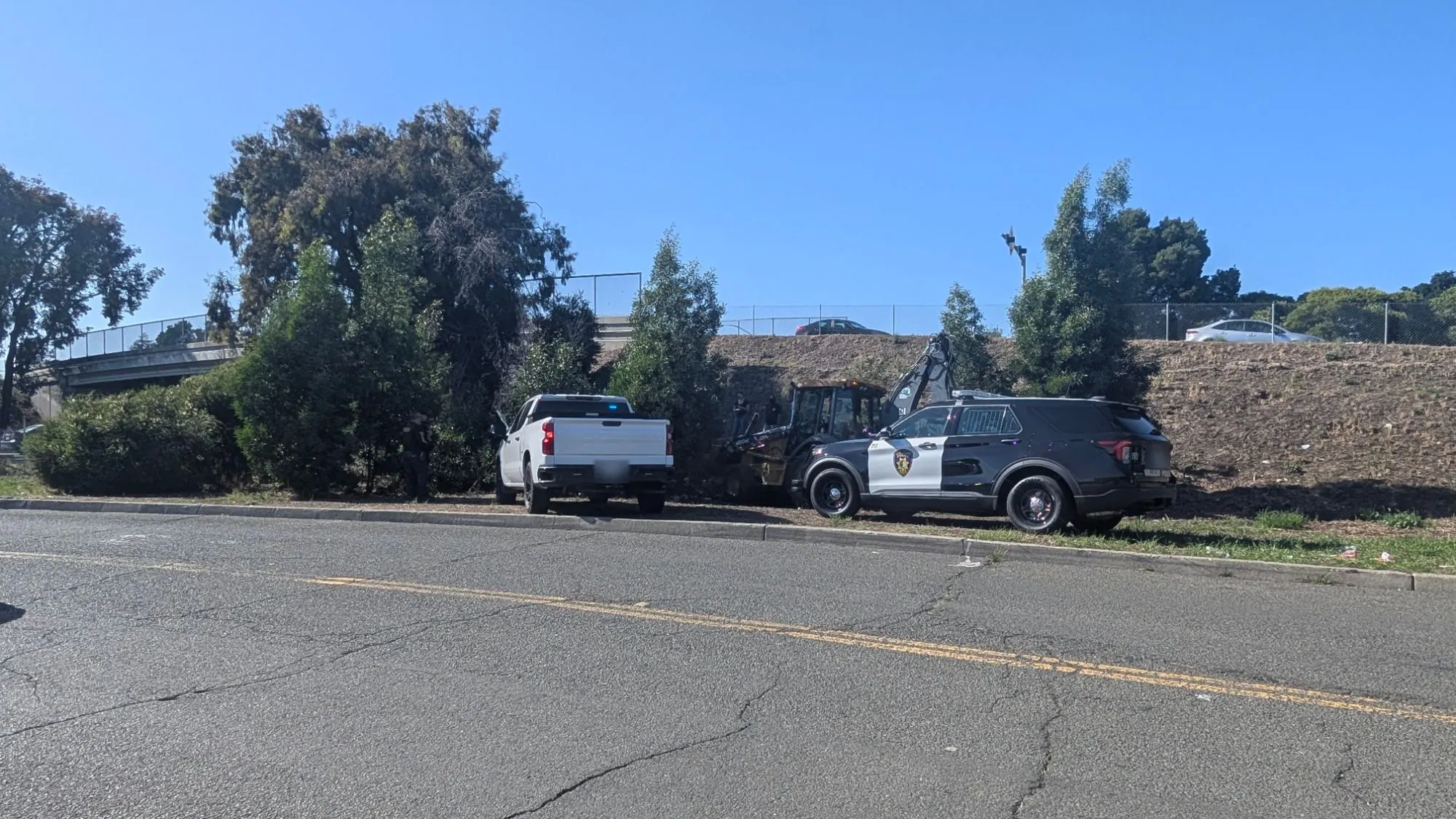 A Vallejo police SUV, a white pickup and an excavator beside a cleared freeway slope in Vallejo, with an officer and two people standing near the trucks